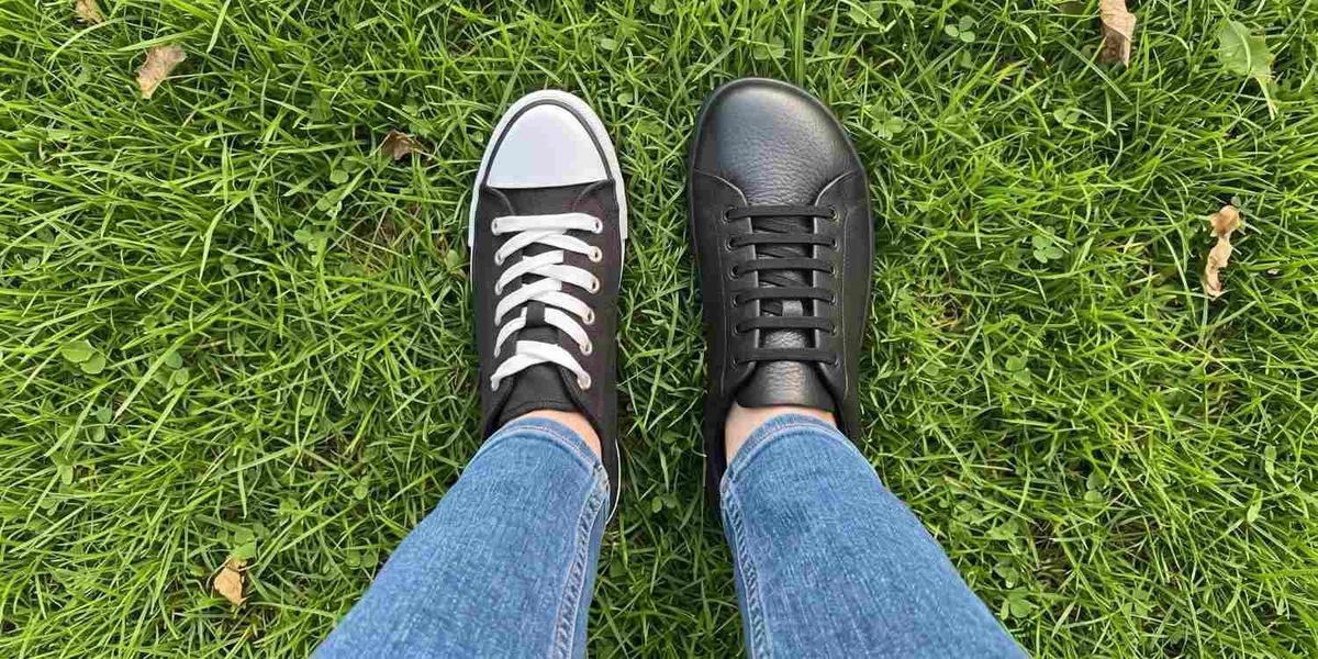 Top-down view of two feet on grass, one in a narrow canvas sneaker and one in a wide, foot-shaped black leather shoe